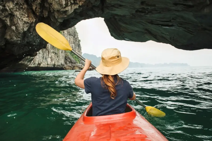 Person kayaking in a red kayak through a scenic sea cave with emerald waters, surrounded by rocky cliffs and distant islands, perfect for adventure and nature tourism.